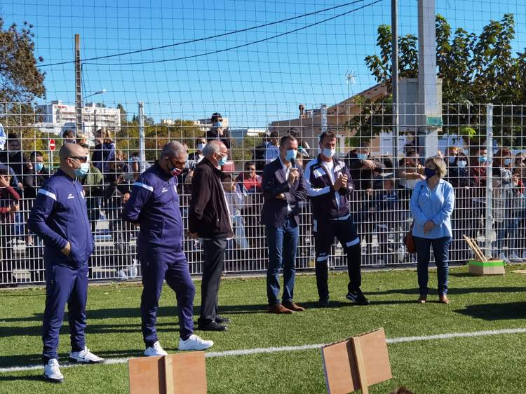 Des officiels et des spectateurs masqués assistent à un événement sur un terrain de football en plein air.