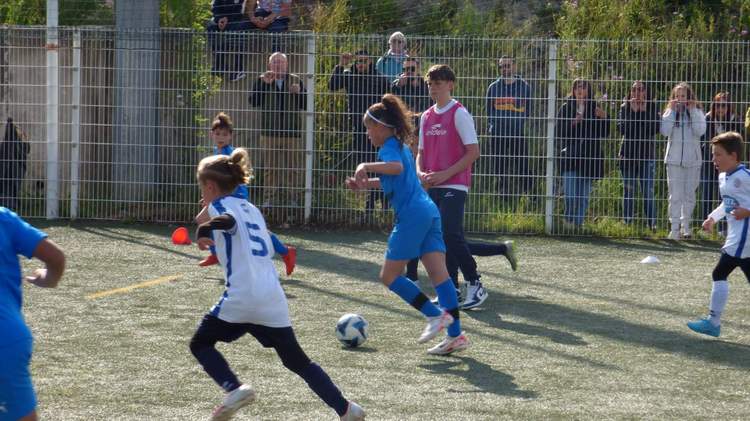 Des jeunes joueurs de football en pleine action sur le terrain, avec des spectateurs derrière une clôture.
