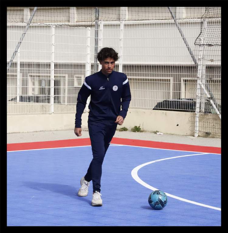 Un jeune joueur en survêtement bleu marine marche sur un terrain de futsal bleu avec un ballon à ses pieds.