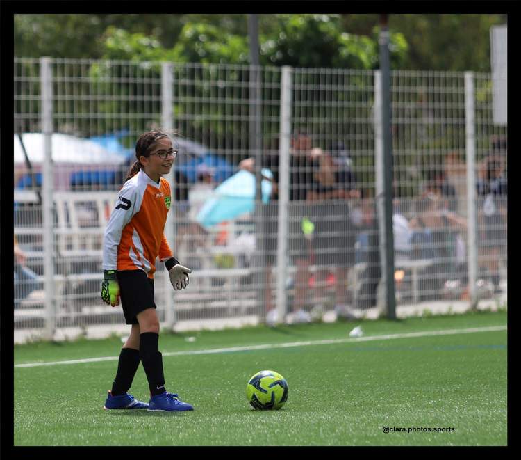 Une jeune gardienne de but en tenue de match, avec des lunettes, se tient sur un terrain de football synthétique à côté d'un ballon.
