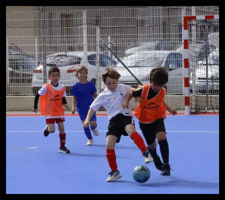 Quatre jeunes joueurs de football en pleine action sur un terrain bleu, avec un but en arrière-plan.